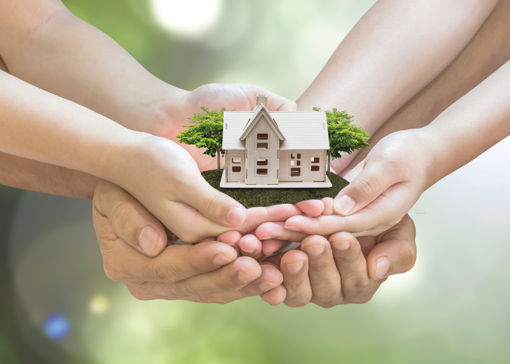 Mehrere Hände halten ein Hausmodell – Symbol für generationsübergreifende Immobilien als Kapitalanlage